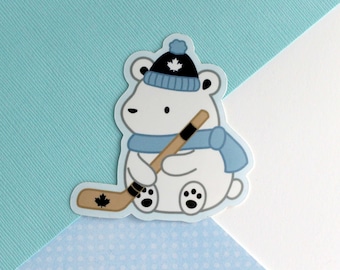 May include: A white polar bear wearing a blue toque with a Canadian maple leaf on it and a blue scarf, holding a hockey stick with a maple leaf on the blade.