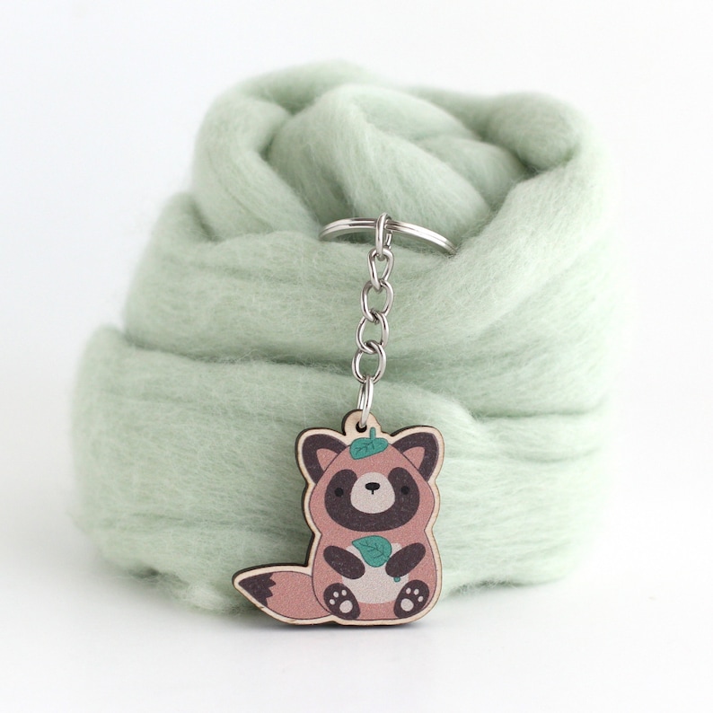 May include: A wooden keychain featuring a cartoon red panda holding a leaf. The keychain is attached to a silver chain and is hanging from a light green fuzzy surface.