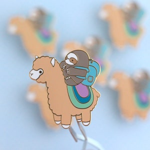 May include: A gold-colored enamel pin featuring a brown sloth wearing a blue backpack riding on a tan alpaca with a rainbow-colored saddle.