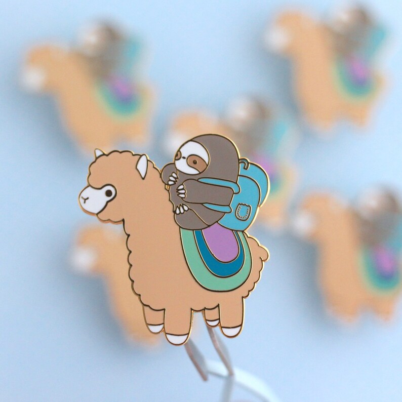 May include: A gold-colored enamel pin featuring a brown sloth wearing a blue backpack riding on a tan alpaca with a rainbow-colored saddle.