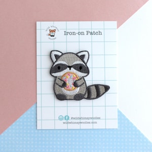 Puede incluir: Un parche termoadhesivo con un mapache de dibujos animados sosteniendo una rosquilla. El mapache es gris, negro y blanco, con una cola a rayas. La rosquilla es rosa, amarilla y azul. El parche est&aacute; en una tarjeta blanca con el texto "Iron-on Patch".