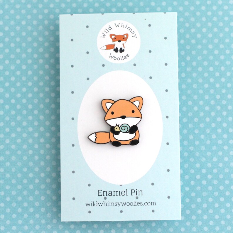 Enamel pin of an cartoon style orange fox holding a little snail, pinned on a backing card
