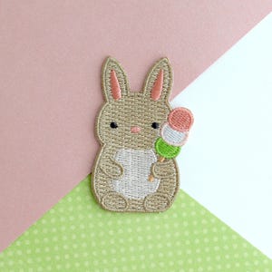May include: Embroidered patch featuring a tan bunny with pink inner ears and a white belly, holding a colorful dango skewer. The patch has a detailed stitched design and is set against a pink and green polka dot background.