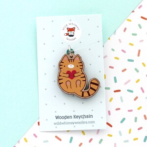 May include: Wooden keychain featuring a cartoon orange tabby cat holding a red heart. The keychain is made by Wild Whimsy Woolies.