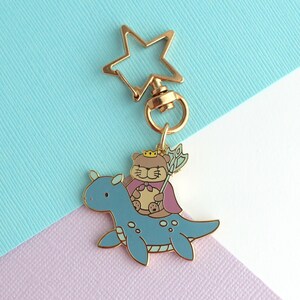 May include: A gold-colored metal keychain featuring a cartoon bear wearing a crown and cape, riding a blue dinosaur-like creature. The keychain has a star-shaped clasp. The bear holds a staff. The background is a mix of blue and purple.