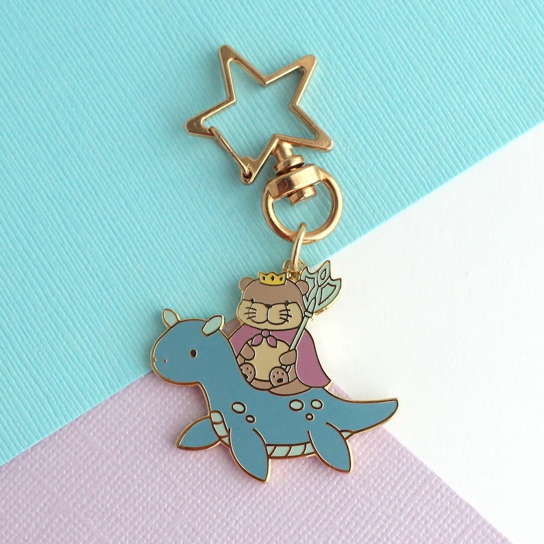 May include: A gold-colored metal keychain featuring a cartoon bear wearing a crown and cape, riding a blue dinosaur-like creature. The keychain has a star-shaped clasp. The bear holds a staff. The background is a mix of blue and purple.