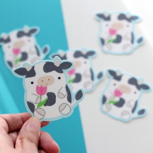 May include: Four black and white cow stickers with pink flowers. The stickers are on a light blue and white background.