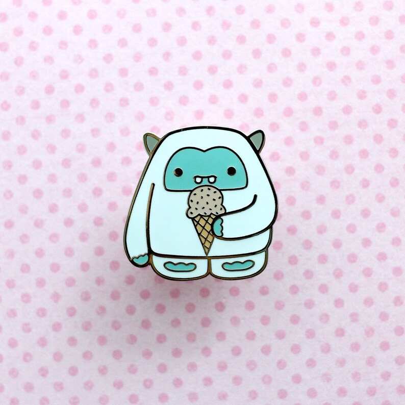May include: A white Yeti with blue eyes and a toothy grin holds a brown and white ice cream cone. The Yeti is wearing a white fur coat and blue booties. The background is a pink polka dot pattern.