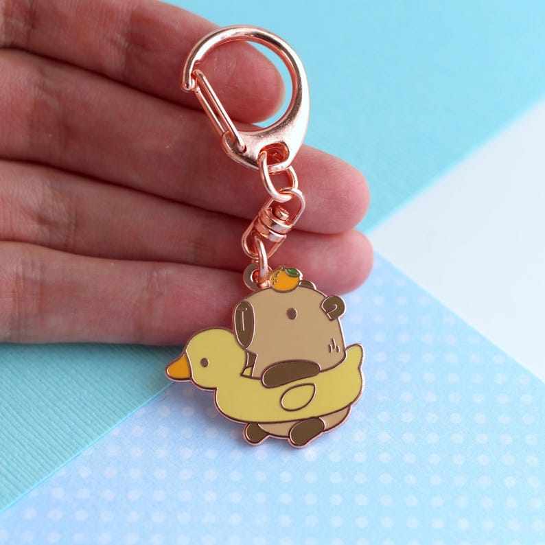 K&ouml;nnte beinhalten: Ein ros&eacute;goldener Schl&uuml;sselanh&auml;nger mit einem Comic-Braunb&auml;ren, der einen gelben Gummiente-Schwimmring mit einer Orangenscheibe auf dem Kopf tr&auml;gt.