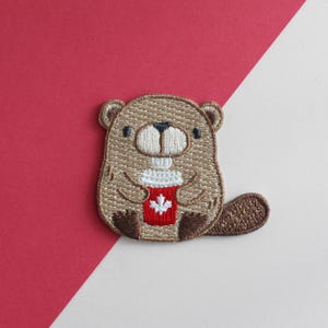 May include: An embroidered patch featuring a light brown beaver with dark brown paws and a tail. The beaver is holding a red cup with a white maple leaf design on the front. The embroidery uses tan, brown, white, black, and red thread colours.