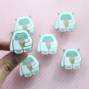 May include: Six white enamel pins with a blue yeti holding an ice cream cone. The yeti has a small mouth and two small eyes. The ice cream cone is brown with a white swirl on top.