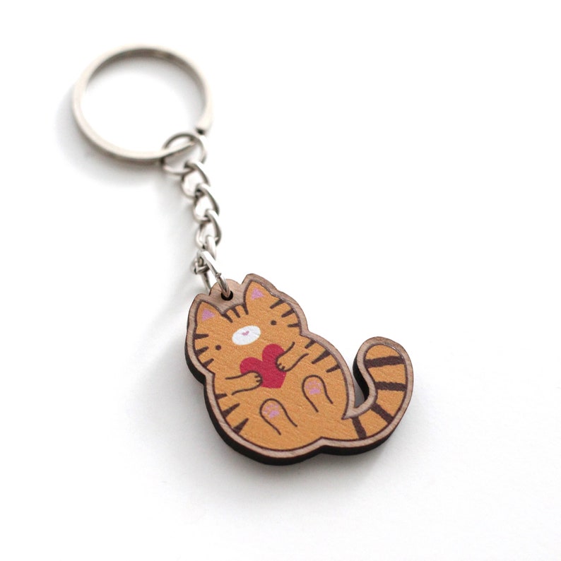 May include: A wooden keychain featuring a cartoon orange tabby cat holding a red heart. The cat has a white face and pink paws.