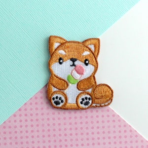 Puede incluir: Parche bordado de un perro Shiba Inu marrón que sostiene un helado de mochi rosa y verde. El perro tiene pelo blanco en el pecho y las patas.