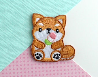 Puede incluir: Parche bordado de un perro Shiba Inu marrón que sostiene un helado de mochi rosa y verde. El perro tiene pelo blanco en el pecho y las patas.