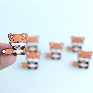 May include: A set of six enamel pins featuring a cartoon fox holding a snail. The pins are orange, brown, and white.