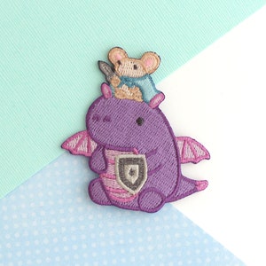 Peut inclure: Patch brodé violet représentant un dragon tenant un bouclier avec une petite souris brune chevauchant sur son dos.