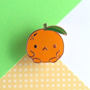 May include: A cute orange enamel pin with a sad face and a green leaf on top. The orange is sitting on a yellow and white polka dot background.