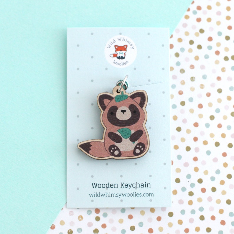 May include: Wooden keychain featuring a cartoon red panda holding a green leaf. The keychain is brown with a light brown tail and white paws. The text "Wooden Keychain" and "wildwhimsywoolies.com" are printed on the card.