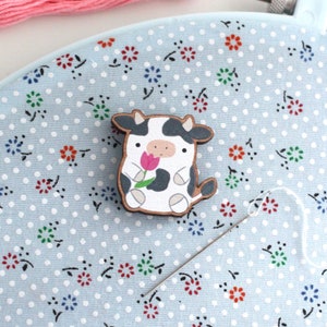 May include: A wooden cow figurine with black and white spots holding a pink flower. The figurine is sitting on a blue and white patterned fabric.