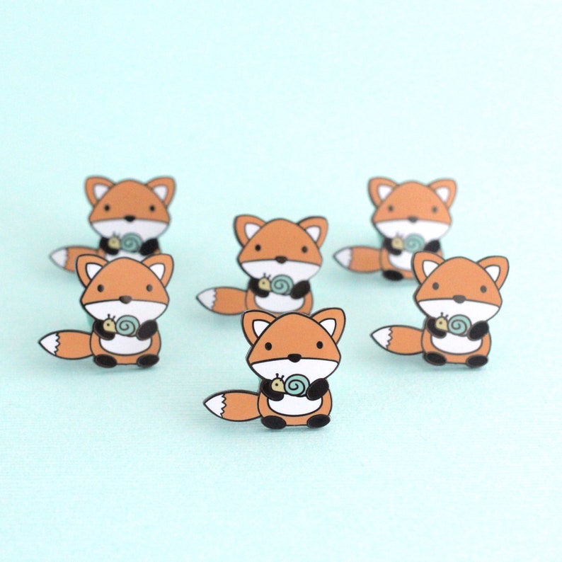Seven enamel pins of a cartoon style orange fox holding a little snail
