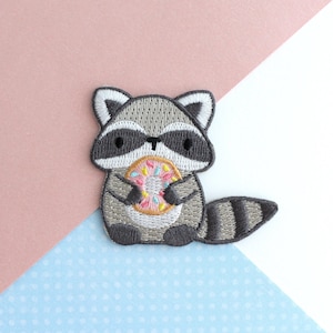 May include: Embroidered patch of a cartoon raccoon holding a donut. The raccoon is grey and white with a black mask and eyes. The donut is pink with colourful sprinkles. The patch is set against a pink and blue patterned background.