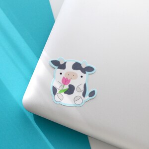 May include: A black and white cow sticker with a pink flower in its mouth. The sticker is on a white laptop.