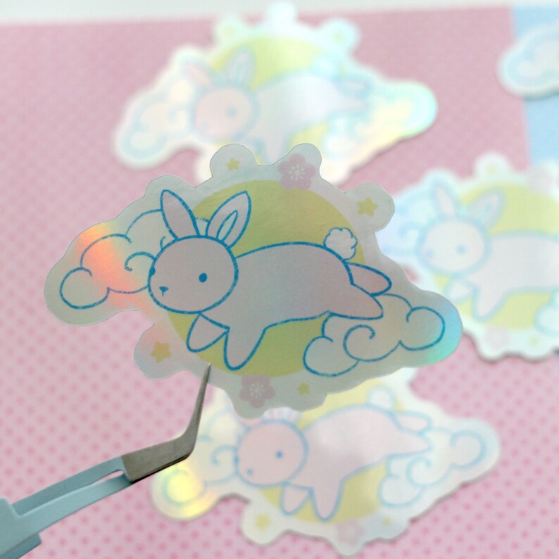 May include: A holographic sticker featuring a cartoon bunny rabbit flying in the clouds with a moon and stars in the background. The sticker is iridescent and has a pastel color scheme.
