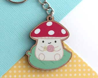 May include: A wooden keychain featuring a cartoon mushroom character with a red cap and white spots. The mushroom is sitting on a green leaf and holding a snail. The keychain has a silver metal chain and ring.