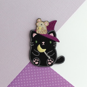 May include: Embroidered patch featuring a black cat wearing a purple witch hat with a small mouse on top. The cat holds a yellow crescent moon wand. The patch has pink accents and green eyes, set against a purple and white background.