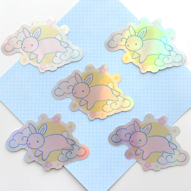 May include: Five iridescent holographic stickers featuring a pink bunny rabbit flying on a cloud with a yellow moon and stars. The stickers are on a light blue background with a white polka dot pattern.