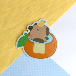 Peut inclure: Un capybara brun portant un canard en caoutchouc jaune prend un bain dans une tranche d'orange remplie d'eau bleue. Le capybara est assis dans la tranche d'orange avec une serviette blanche enroulée autour de son corps.
