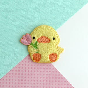 May include: Embroidered yellow duck patch holding a pink tulip. The duck has orange feet and a matching beak. The patch is on a background of light blue, white, and pink polka dots.