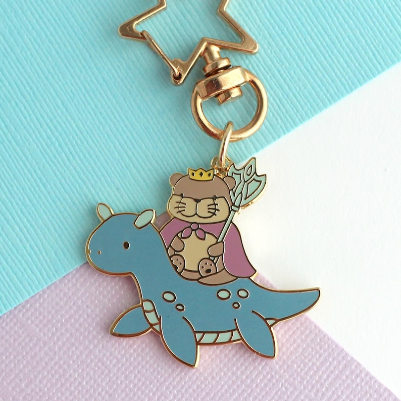 May include: A gold-colored keychain featuring a cartoon bear wearing a crown and cape, riding a blue dinosaur-like creature. The bear holds a scepter. The keychain has a star-shaped clasp.