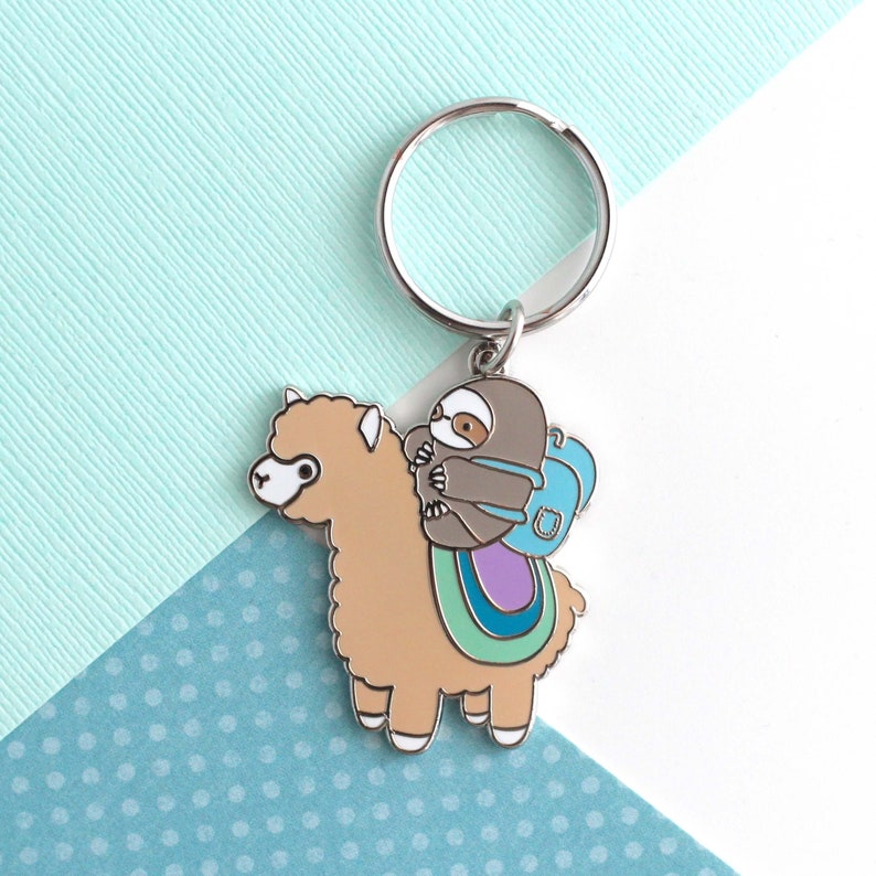 Silver plated keychain of little grey sloths wearing a backpack and riding on top of a beige alpaca (or llama)