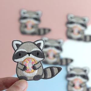 Puede incluir: Parche bordado con un mapache de dibujos animados sosteniendo una rosquilla. El mapache tiene pelaje gris y blanco, una m&aacute;scara negra y una cola a rayas. La rosquilla es rosa, amarilla y azul. Se ven otros parches de mapaches en el fondo.