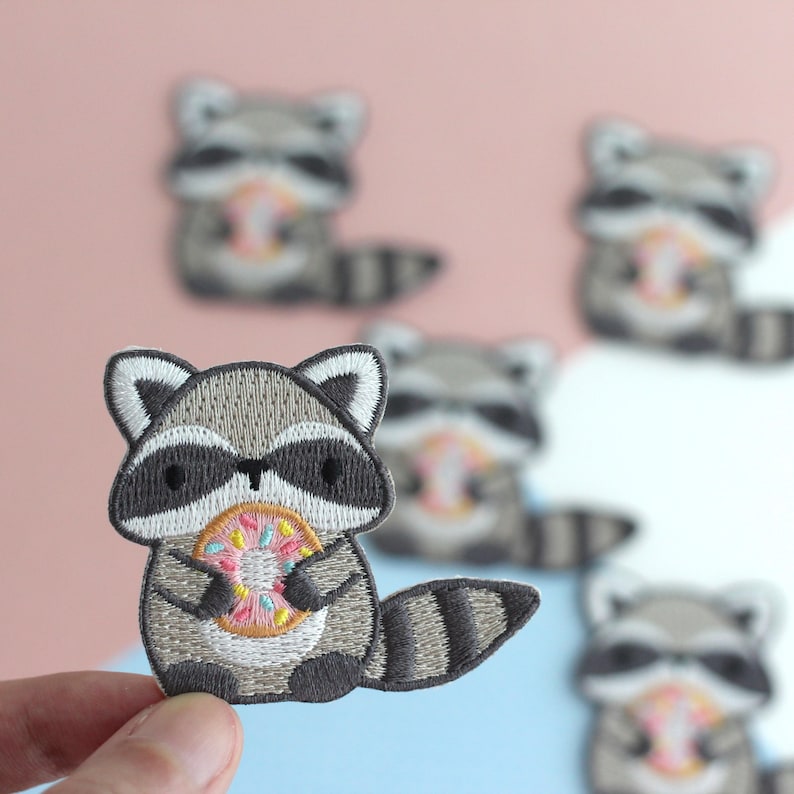 Puede incluir: Parche bordado con un mapache de dibujos animados sosteniendo una rosquilla. El mapache tiene pelaje gris y blanco, una m&aacute;scara negra y una cola a rayas. La rosquilla es rosa, amarilla y azul. Se ven otros parches de mapaches en el fondo.