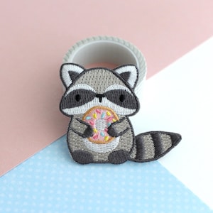 Puede incluir: Parche bordado con un mapache de dibujos animados sosteniendo una rosquilla. El mapache tiene pelaje gris y blanco con ojos negros y una cola a rayas. La rosquilla es rosa con chispas de colores. Para ropa o accesorios.