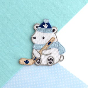 May include: Embroidered patch of a white polar bear wearing a blue hat and scarf, holding a hockey stick. The bear has black eyes and paws. The hockey stick and hat feature a maple leaf design.