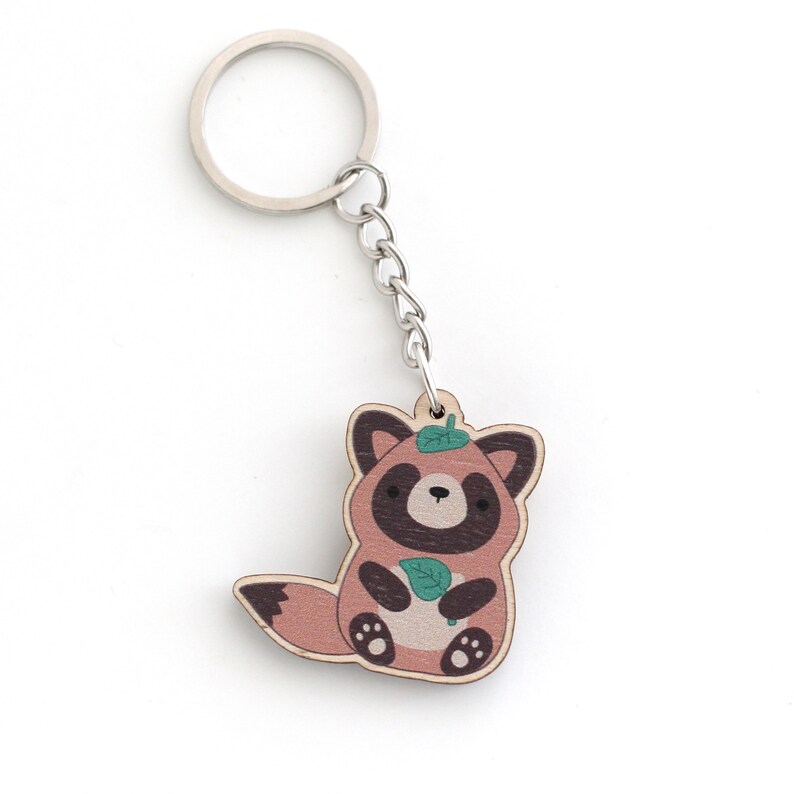 May include: A wooden keychain featuring a cartoon raccoon holding a green leaf. The raccoon is brown with a black face and has a green leaf on its head. The keychain is attached to a silver metal ring.
