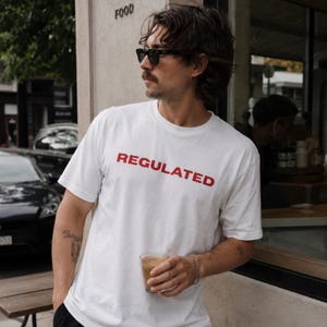 May include: A white short-sleeved t-shirt featuring the word REGULATED printed in bold red capital letters across the chest. The garment has a classic crew neckline and a straight hem, constructed from a plain white fabric with a simple design.