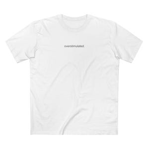 May include: A white short-sleeved crew neck t-shirt featuring the word overstimulated. printed in small, centred black lowercase letters on the upper chest area. The garment is made of a plain white fabric with a standard fit and short sleeves.