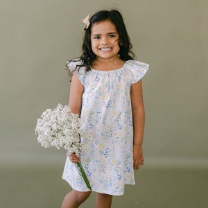 May include: A young girl in a white dress with a floral pattern holds a bouquet of white baby's breath flowers. She is smiling and looking at the camera.