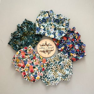 May include: Six pairs of colorful floral print baby bloomers arranged in a circle. The bloomers are made of soft cotton fabric and have a ruffled elastic waistband. The bloomers are in a variety of colors, including blue, green, red, and yellow. The bloomers are arranged in a circle around a wooden sign that says "willow + hazel handmade".