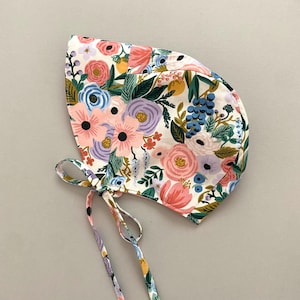 May include: A floral print baby bonnet with a white background. The bonnet features a colorful pattern of pink, blue, purple, and yellow flowers with green leaves. The bonnet has ties on each side.