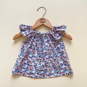 May include: A white, blue, and pink floral print baby top with ruffled sleeves. The top is hanging on a wooden hanger with the text "willow & bay" printed on it.