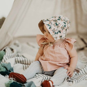 K&ouml;nnte beinhalten: Ein Baby tr&auml;gt eine Bl&uuml;mchenm&uuml;tze, ein rosa Oberteil und wei&szlig;e Strumpfhosen und sitzt auf einer gestreiften Decke. Das Baby tr&auml;gt braune Schuhe.