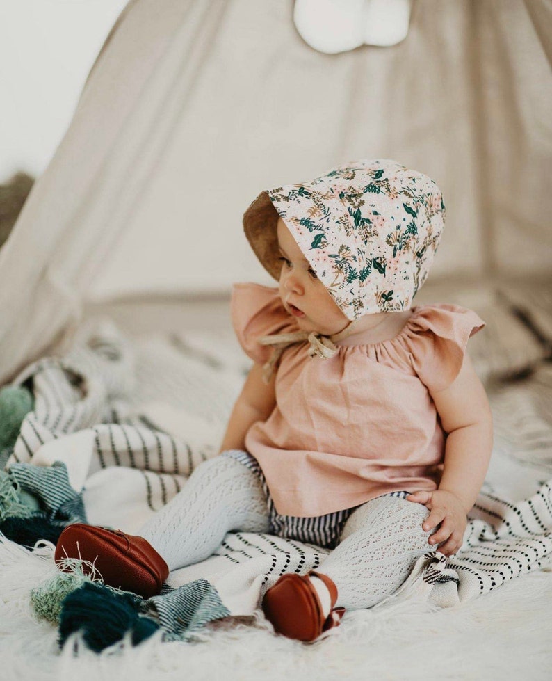 K&ouml;nnte beinhalten: Ein Baby tr&auml;gt eine Bl&uuml;mchenm&uuml;tze, ein rosa Oberteil und wei&szlig;e Strumpfhosen und sitzt auf einer gestreiften Decke. Das Baby tr&auml;gt braune Schuhe.