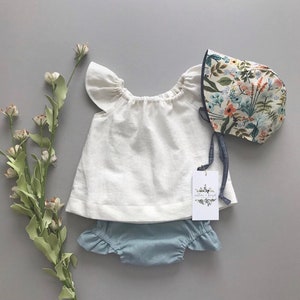 May include: A white linen baby top with ruffled sleeves, blue denim bloomers with ruffles, and a floral print sunbonnet with a blue ribbon.