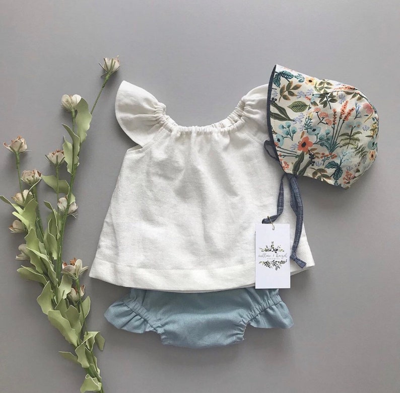 May include: A white linen baby top with ruffled sleeves, blue denim bloomers with ruffles, and a floral print sunbonnet with a blue ribbon.