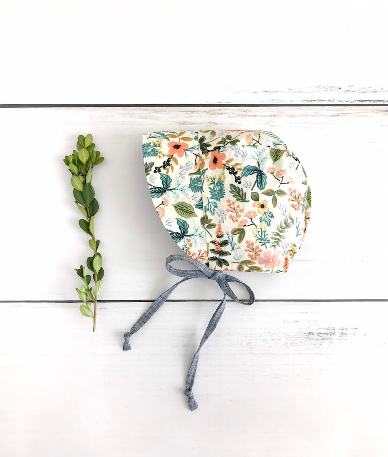 May include: A white bonnet with a floral print featuring pink, orange, and green flowers. The bonnet has a blue ribbon tie.